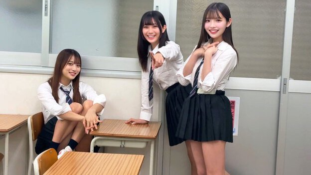 教室で女子3人が机の前に集まり、スカート姿で笑いながら男子へ視線を向ける